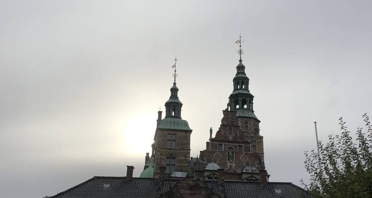 Les deux flèches élancées du château de Rosenborg en silhouette avec un soleil voilé derrière un ciel gris.