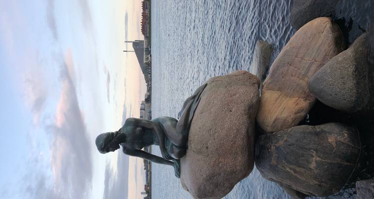 Statue de bronze iconique de la Petite Sirène perchée sur des rochers de granit dominant le port au crépuscule.