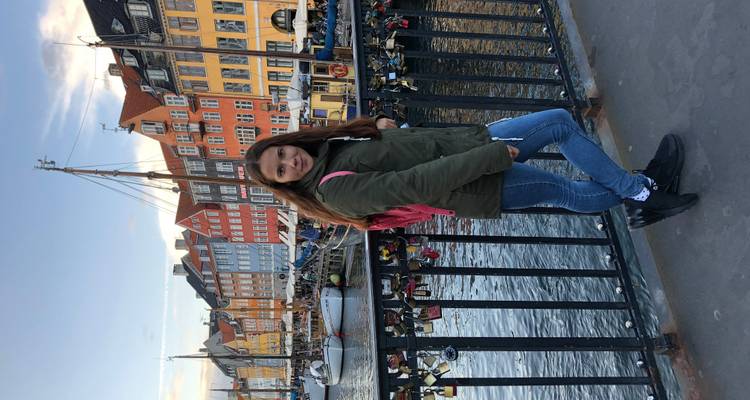 Une jeune femme pose sur un pont du canal à Nyhavn avec des maisons historiques colorées et des bateaux derrière elle.