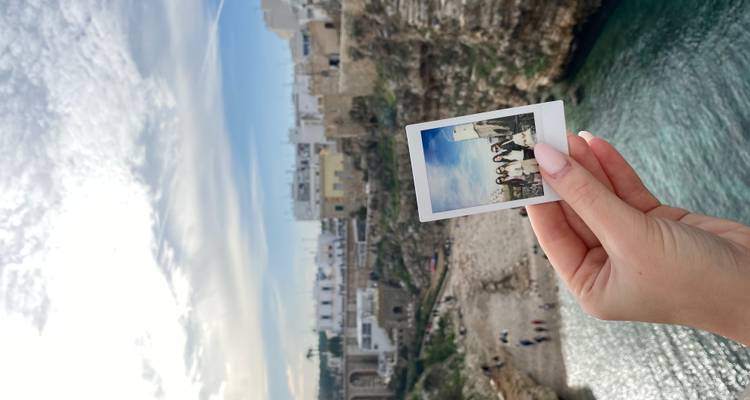 Hand houdt een polaroidfoto tegen de kliffstad en blauwe zee van Polignano a Mare.