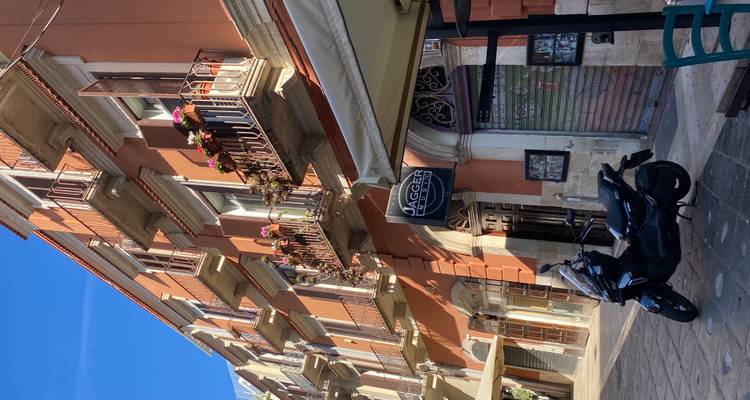 Een zonnige Italiaanse stadswoning met balkons en een scooter geparkeerd op een rustige straat.
