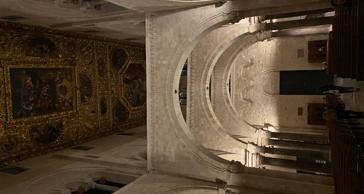 Zwak verlichte binnenkant van een stenen gewelfdе kathedraal met een sierlijk beschilderd plafond en bezoekers die door het middenschip wandelen.