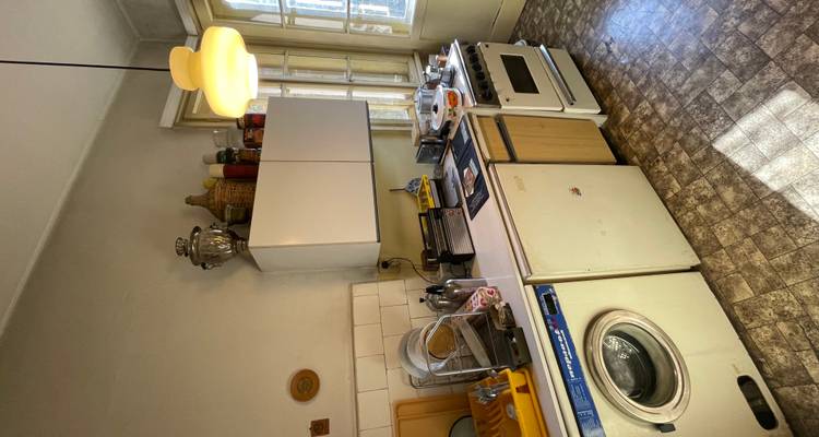 Cocina vintage con electrodomésticos antiguos, lavadora y estantes desordenados bañados por la luz del día.