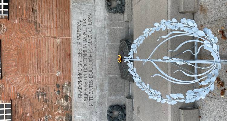 Muro conmemorativo de piedra con inscripción en búlgaro y una escultura de corona con llama eterna.