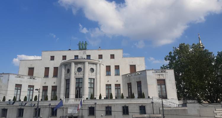 Edificio de estilo palacio de mármol blanco con banderas ondeando al frente, recortado contra un cielo azul brillante.