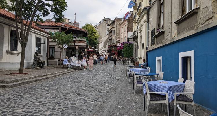 Callejón estrecho y empinado de adoquines en Skadarlija con mesas al aire libre cubiertas de telas azules y turistas paseando.