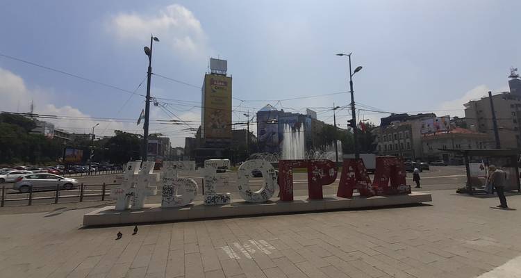 Gran letrero #BELGRADO y fuente en una plaza urbana concurrida con tráfico y vallas publicitarias.
