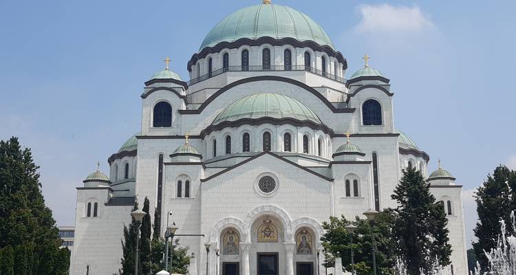 Fachada frontal de la monumental Iglesia blanca de San Sava con múltiples cúpulas verdes e iconos.