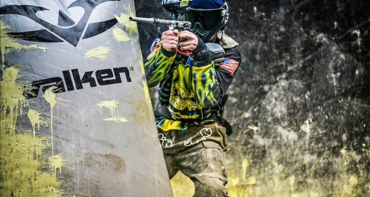 Aktionsfoto eines Paintball-Spielers, der sein Marker hinter einer mit Spritzern bedeckten Barriere abfeuert.