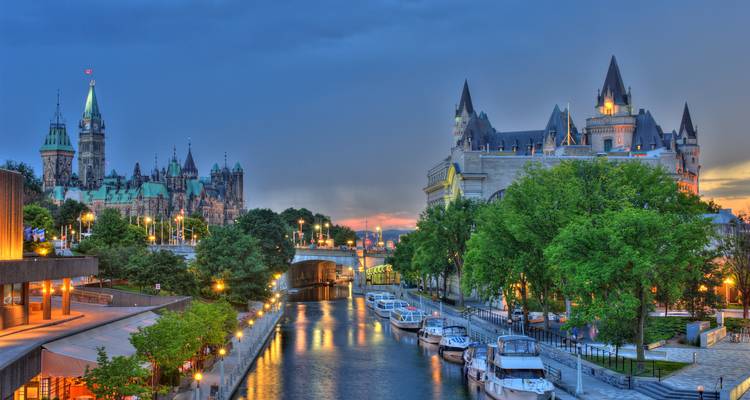 Vue en soirée des édifices du Parlement d'Ottawa et du Château Laurier surplombant le canal Rideau illuminé et les bateaux amarrés.