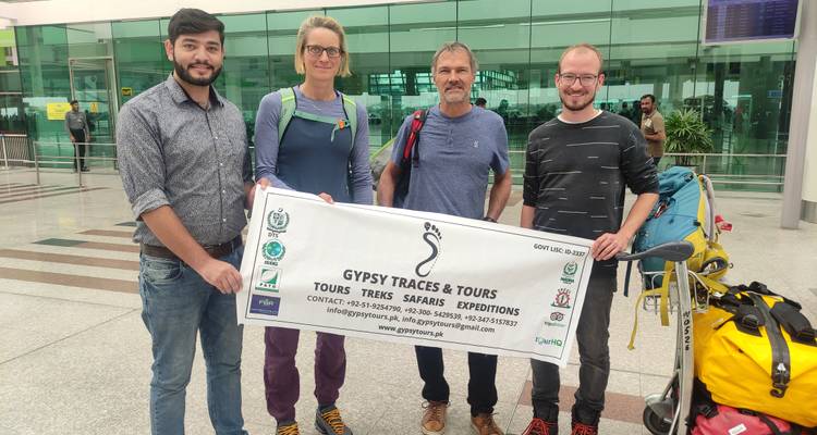 Cuatro viajeros posan dentro de una terminal de aeropuerto moderna sosteniendo un banner de tour con equipo de trekking a sus pies.