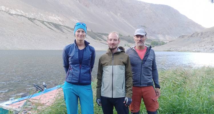 Tres trekkers están de pie junto a un lago de montaña remoto rodeado de pendientes rocosas desoladas y pastos altos.