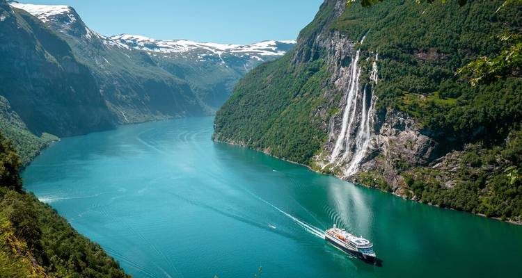 Un navire de croisière glisse à travers le Geirangerfjord turquoise en passant devant une haute cascade et des pics enneigés.