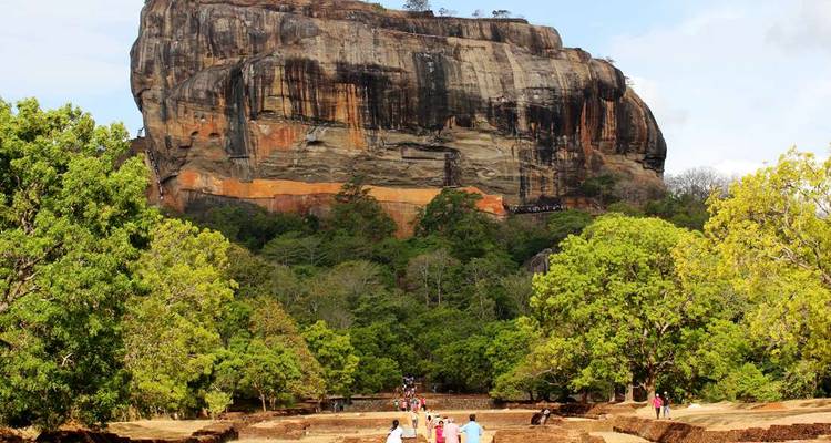 Toeristen wandelen over het oude terrein onder de uitstekende Sigiriya Rock Fortress die boven de jungle uittorent.