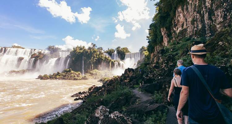 Besucher folgen einem üppigen Wanderweg neben den donnernden Wasserfällen der Iguazu-Wasserfälle an einem sonnigen Tag.