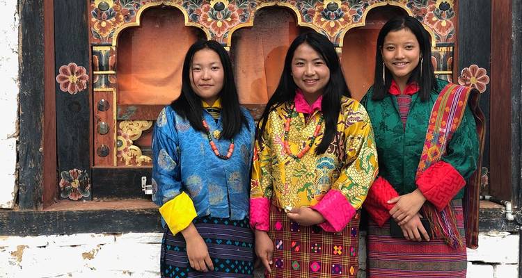 Trois jeunes femmes bhoutanaises vêtues de kira traditionnels colorés se tiennent souriantes devant une fenêtre de temple richement décorée.