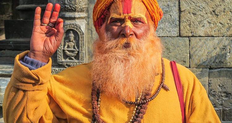 Un sadhu peint de couleurs vives, avec une barbe orange et une robe jaune, bénit l'appareil photo de sa main levée contre un mur de temple en pierre.