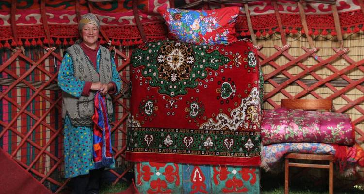 Oudere Kirgizische vrouw in een gedessineerde jurk staat in een rijk versierde yurt vol kleurrijke textielwerk en tapijten.
