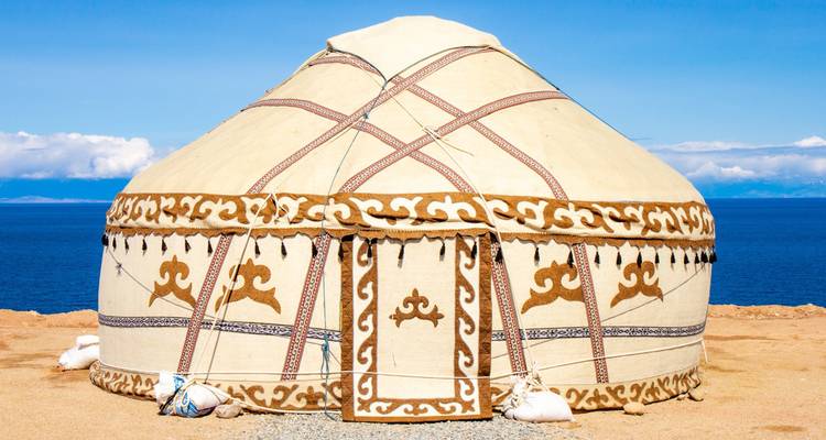 Una yurta kirguís tradicional ubicada en una meseta de orilla de lago azotada por los vientos, con agua y cielo de un azul profundo más allá.