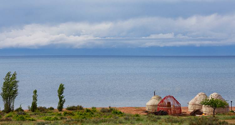 Grupo de yurtas y una estructura de madera junto a la tranquila extensión azul del lago Issyk Kul bajo un cielo nublado.