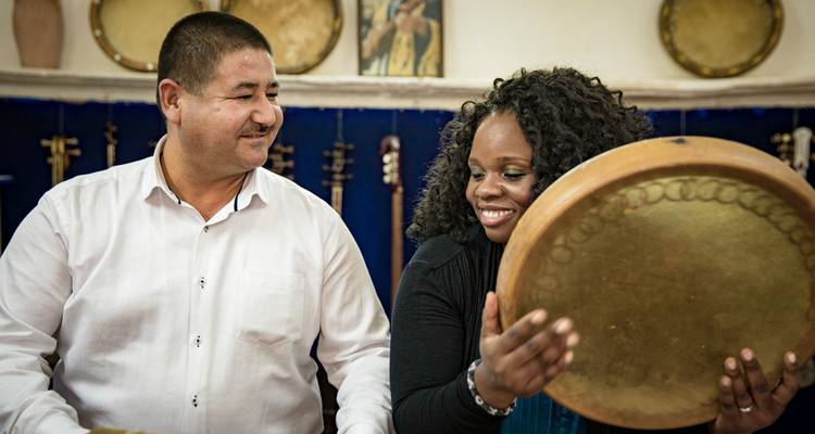 Un homme et une femme souriants expérimentent un grand tambour sur cadre ouzbek à l'intérieur d'un atelier musical doté d'instruments.