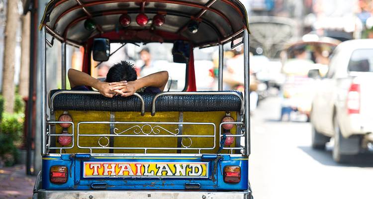 Un homme se détend, les bras croisés derrière la tête, à l'intérieur d'un tuk-tuk aux couleurs vives, dans une rue thaïlandaise animée.
