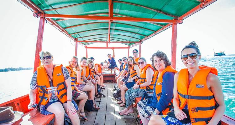 Groupe de touristes souriants vêtus de gilets de sauvetage orange assis sur un bateau en bois couvert glissant sur une eau turquoise