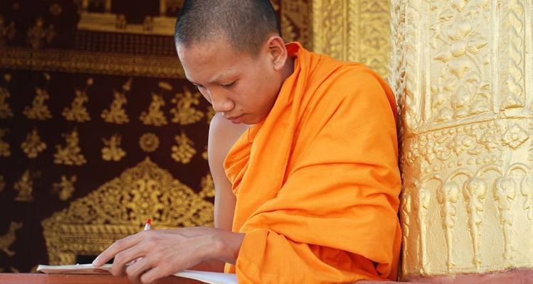 Jeune moine bouddhiste en robes orange se concentrant sur l'écriture près d'un pilier doré richement sculpté