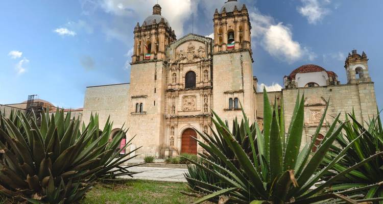 Ornate stone facade of Santo Domingo de Guzmán church framed by agave plants and blue sky