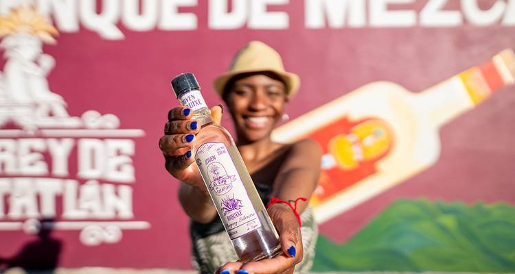 Smiling traveller proudly holds out a bottle of mezcal against a colorful mural backdrop