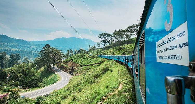 Bright blue passenger train curling through lush green tea-covered hills of Sri Lanka with mountains in the background.