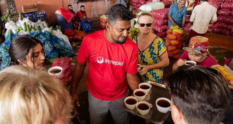 Travellers sampling cups of tea in a busy produce market surrounded by sacks of vegetables and fruit.