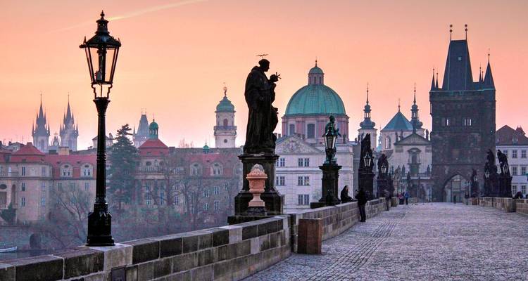 Die Karlsbrücke bei Sonnenaufgang mit Statuen, die sich gegen einen Pastellhimmel über Prags historischer Skyline abzeichnen.