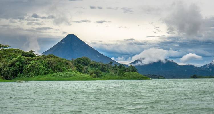 Conical Arenal Volcano rising above Lake Arenal with lush rainforest and drifting clouds.