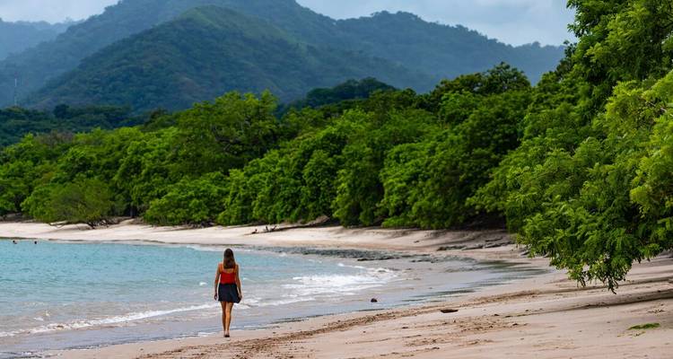 Woman strolling along a quiet tropical beach with jungle-covered hills in the background.