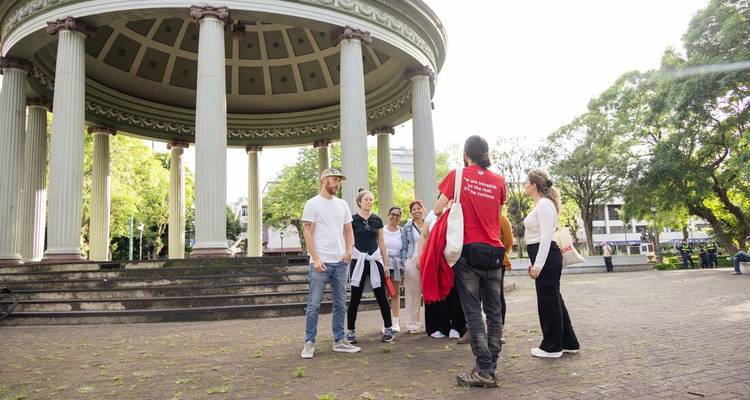 Travellers listen to a guide beside a classical gazebo with tall columns in a leafy city park.