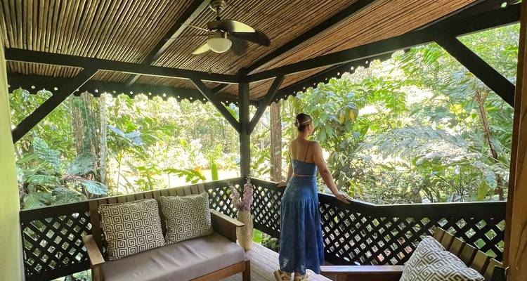 Woman in a blue dress stands on a wooden jungle balcony overlooking lush green foliage.
