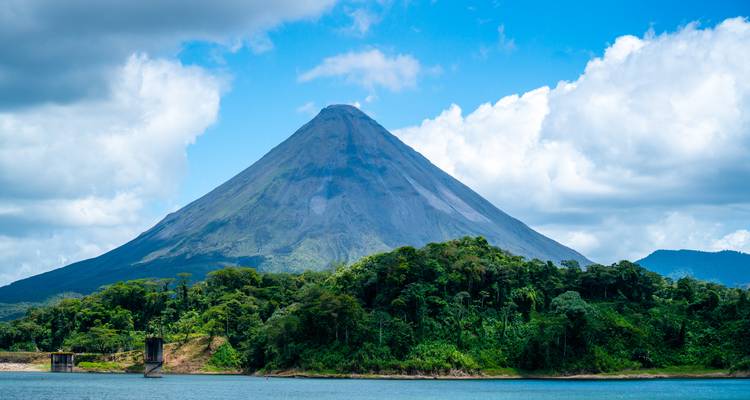 El cono perfecto del Volcán Arenal se alza sobre la exuberante selva tropical y un lago azul bajo nubes dispersas.