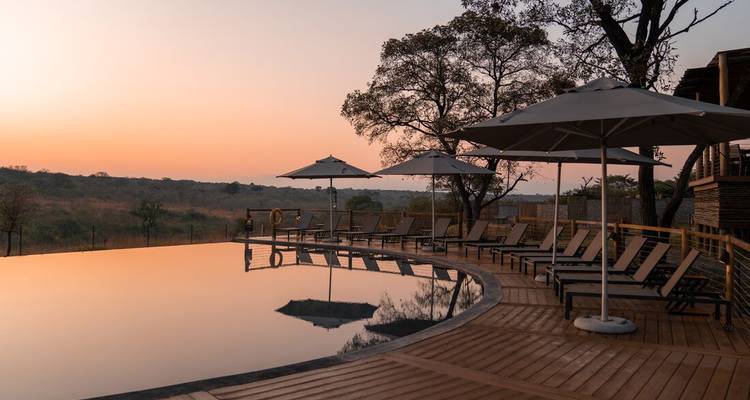 An infinity pool lined with lounge chairs and umbrellas reflects the pastel colors of sunset over distant hills.
