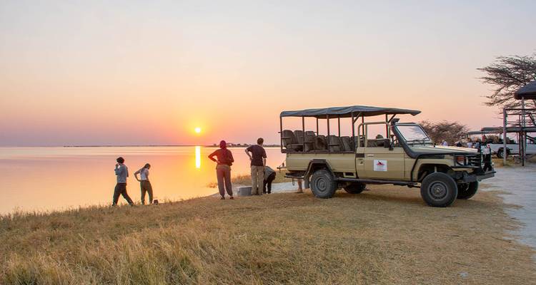 Tourists stand by a safari truck sipping drinks while watching the sun set over a calm lake.