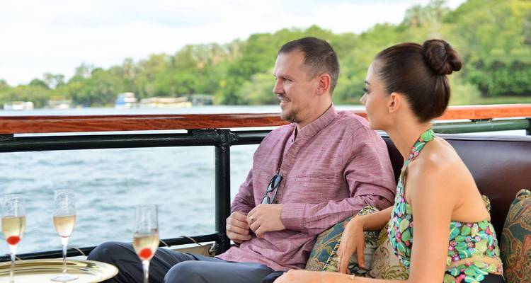 A couple relaxes with champagne on the deck of a river cruise boat, lush banks in the background.