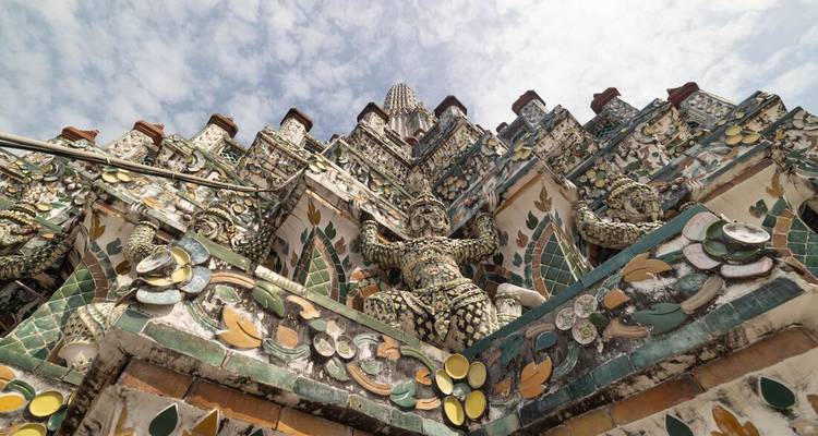 Détails céramiques et floraux complexes du temple Wat Arun s'élevant vers un ciel partiellement nuageux.