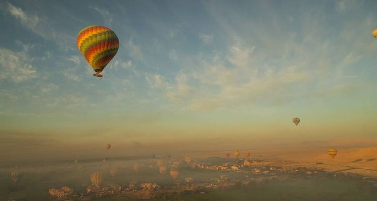 Colorful hot-air balloons float above a hazy Nile Valley landscape at dawn.