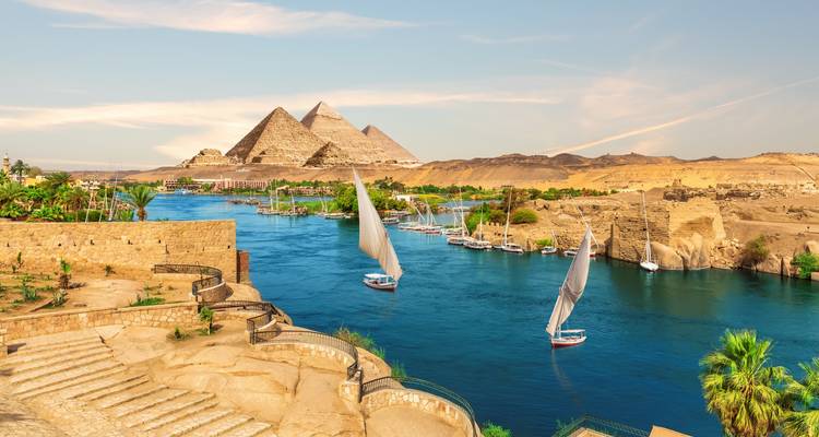 An idyllic scene of the Nile River with feluccas sailing past the three Giza pyramids rising from the desert.