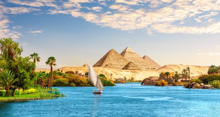 Bright blue Nile waters bordered by lush palms lead the eye to the iconic Giza pyramids under a lightly clouded sky.