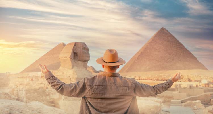 A traveller with outstretched arms admires the Sphinx and pyramids bathed in warm evening light.