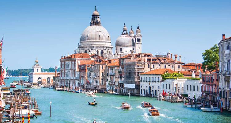 Des bateaux naviguent le long du Grand Canal de Venise en direction des dômes de la Basilique Santa Maria della Salute sous un ciel bleu dégagé.