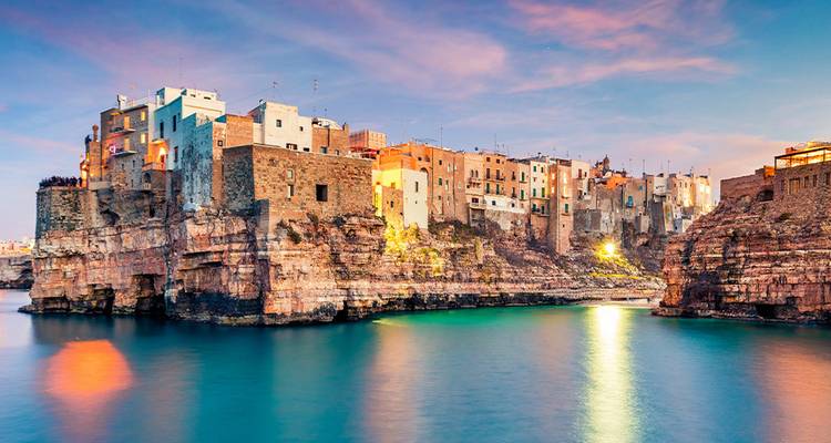 Des maisons colorées en bord de mer se perchent au sommet de falaises escarpées surplombant des eaux turquoise calmes au crépuscule.