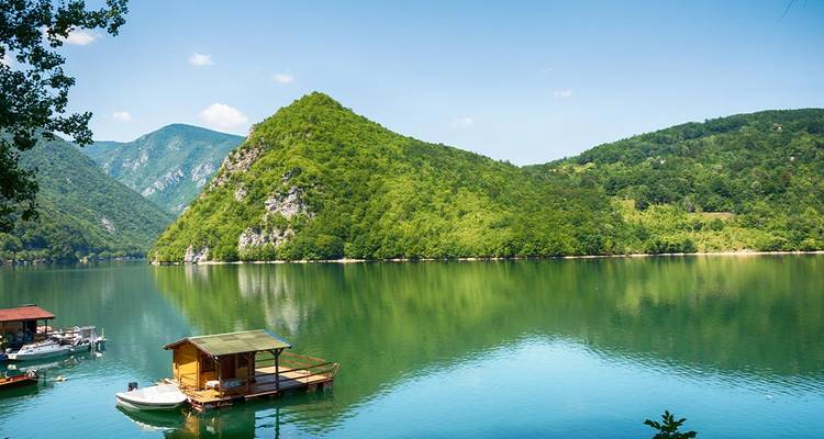 Calm emerald lake reflecting lush green mountains and a small floating cottage.