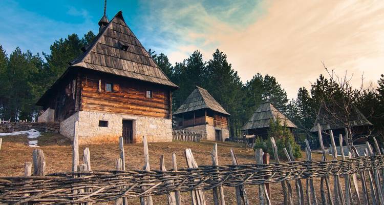 Traditional wooden cottages with shingled roofs behind a woven fence at an open-air museum.